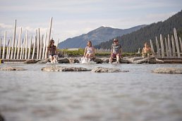 Familie sitzt gemeinsam am Bach und hält die Füße ins Wasser